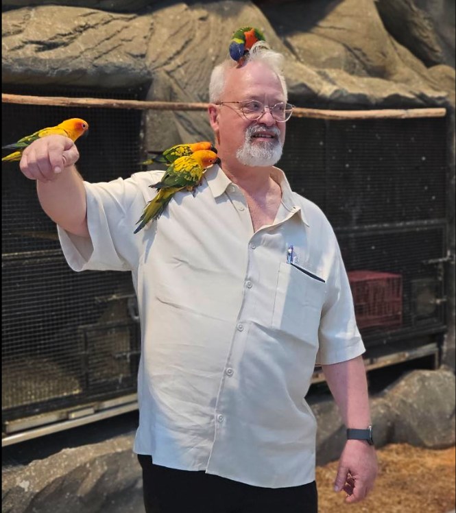 Dr. David P. Smith smiling in an aviary with colorful parrots perched on his arm, shoulder, and head
